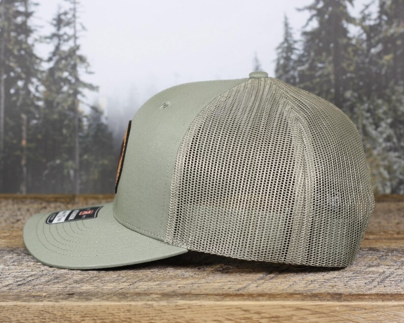 May include: A sage green baseball cap with a mesh back and a curved brim. The cap features a rectangular patch with a design and a small label on the brim. The hat is displayed on a wooden surface.
