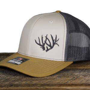 May include: A beige and gray trucker hat with a curved brim. The front panel features an embroidered antler design in dark gray. The brim is tan, and the mesh back is gray. The hat has a snapback closure.