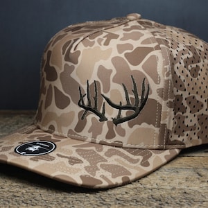 May include: A brown and tan camouflage trucker hat with a black embroidered deer antler design. The hat has a mesh back.