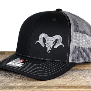 May include: Black and gray trucker hat with a ram skull embroidered on the front. The hat features a curved brim, a mesh back, and an adjustable snap closure. The Richardson brand label is visible on the front.