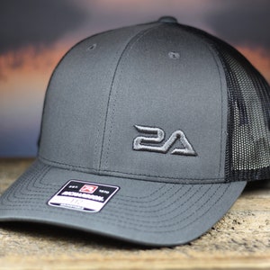May include: A charcoal gray trucker hat with a black mesh back. The hat features a curved brim and a stitched logo on the front. A small tag is visible on the brim.
