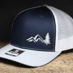 May include: A navy blue and white trucker hat with a white embroidered design of mountains and a pine tree. The hat has a gray undervisor and a mesh back.