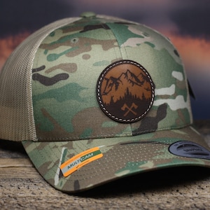 May include: A camouflage trucker hat with a brown leather patch featuring a mountain scene and crossed axes. The patch is stitched around the edges. The hat has a mesh back and a "MULTICAM" label on the side.