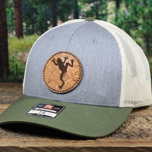 May include: A gray trucker hat with a green brim and a white mesh back. The hat features a circular leather patch with a frog design. The patch is brown with a topographic map design. The hat has a Richardson 115 tag.