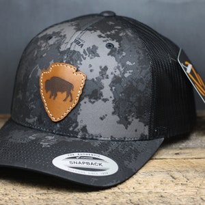 May include: Black and gray camouflage trucker hat with a brown leather patch featuring a bison silhouette and the text "The Authentic Snapback" on the underside of the brim.
