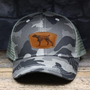 May include: A camouflage trucker hat with a brown leather patch featuring a black and white silhouette of a dog.