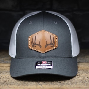 May include: A gray and white trucker hat with a brown leather patch featuring a deer antler design. The hat has a mesh back and a Richardson 115 label.