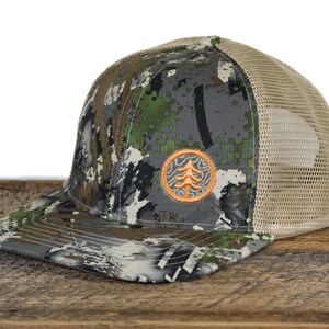 May include: A camouflage baseball cap with a tan mesh back and a flat brim. The front panel features a circular orange embroidered patch with a stylized tree design. The camouflage pattern consists of green, brown, black, and white pixelated shapes.