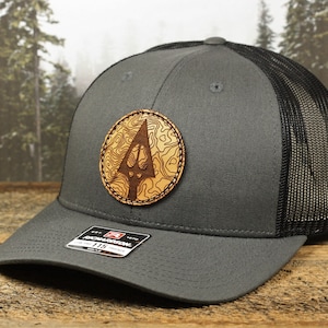 May include: A gray trucker hat with a black mesh back and a curved brim. The hat features a circular, wooden patch with a topographic map design and an arrow. The patch is centered on the front of the hat.