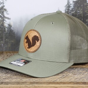 May include: A sage green trucker hat with a brown leather patch featuring a squirrel silhouette. The hat has a mesh back and a curved brim. The patch has a topographic design.