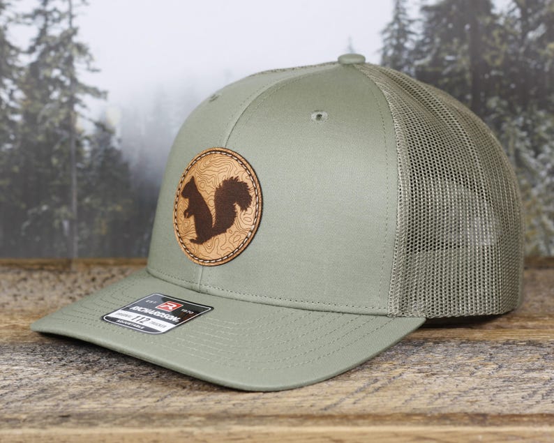 May include: A sage green trucker hat with a brown leather patch featuring a squirrel silhouette. The hat has a mesh back and a curved brim. The patch has a topographic design.