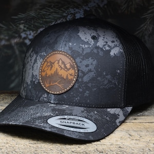 May include: A black and gray camouflage trucker hat with a brown leather patch featuring a mountain scene. The hat has a black mesh back and a snap closure. The text "The Authentic Snapback" is printed on the closure.