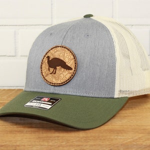 May include: A gray and green trucker hat with a brown leather patch featuring a silhouette of a wild turkey. The patch has a topographic design.