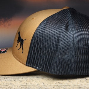 May include: A brown and black trucker hat with a black silhouette of a dog on the front. The hat has a mesh back.