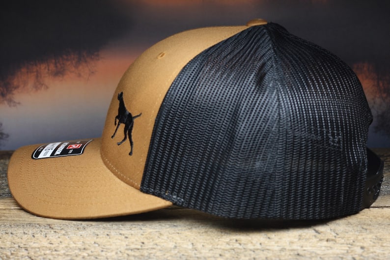 May include: A brown and black trucker hat with a black silhouette of a dog on the front. The hat has a mesh back.