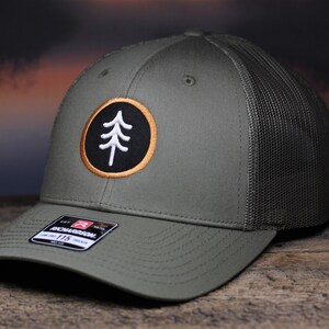 May include: Olive green trucker hat with a black mesh back. The front features a circular patch with a white tree design on a black background, framed by a gold border. The hat has a curved brim and a Richardson 115 tag.