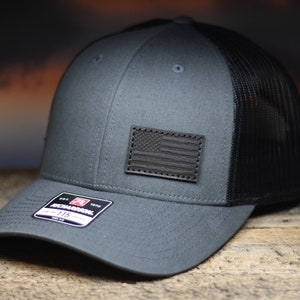 May include: A charcoal gray trucker hat with a black mesh back and a curved brim. A black leather patch with an American flag design is on the front. The hat has a Richardson 115 tag.
