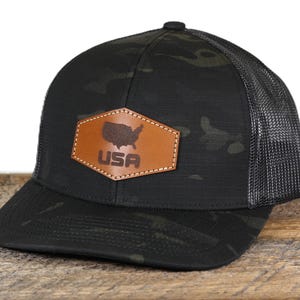 May include: Black camouflage baseball cap with a mesh back and a curved brim. The cap features a brown leather patch with the outline of the United States and the word "USA" in bold letters.