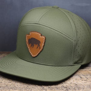 May include: A green, breathable,  mesh-backed baseball cap with a brown leather patch featuring a silhouette of a buffalo. The patch is shaped like a shield with a white stitching outline.