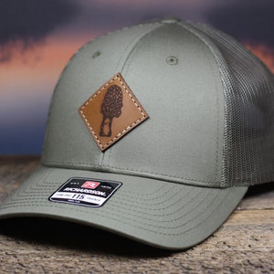 May include: Olive green trucker hat with a brown leather patch featuring a mushroom design. The hat has a mesh back and a curved brim. The Richardson 115 Trucker hat is a low-profile style.