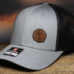 May include: Gray and black trucker hat with a tree ring patch. The hat features a gray front panel and a black mesh back. A small patch with a tree ring design is sewn on the front. The hat has a curved brim and a tag that reads "Richardson 115 Trucker".