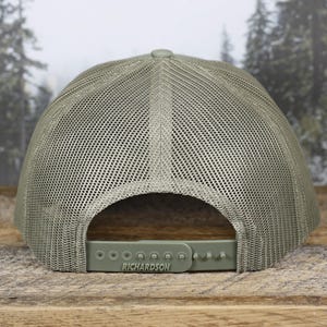 May include: A sage green trucker hat with a mesh back and a curved brim. The adjustable snap closure features the brand name "RICHARDSON". The hat is displayed on a wooden surface with a blurred forest background.