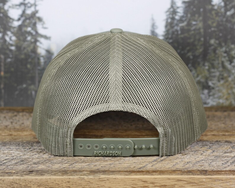 May include: A sage green trucker hat with a mesh back and a curved brim. The adjustable snap closure features the brand name "RICHARDSON". The hat is displayed on a wooden surface with a blurred forest background.