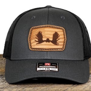 May include: A charcoal gray trucker hat with a black mesh back. The front features a rectangular leather patch with a moose silhouette over a faded American flag design. The hat has a curved brim and a Richardson 115 Trucker tag.