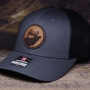 May include: A gray and black trucker hat with a brown leather patch featuring a silhouette of an elk with antlers. The patch has a topographic design around the elk. The hat has a black mesh back and a Richardson 115 label.