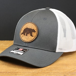 May include: A gray and white trucker hat with a brown leather patch featuring a silhouette of a bear. The patch has a topographic design.