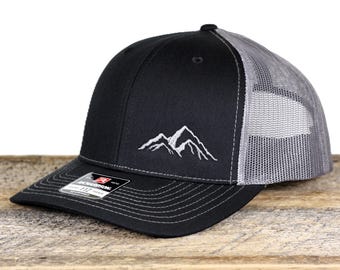 May include: Black baseball cap with a grey mesh back and a white embroidered mountain range design. The cap has a curved brim and a snapback closure. The hat is sitting on a wooden surface.