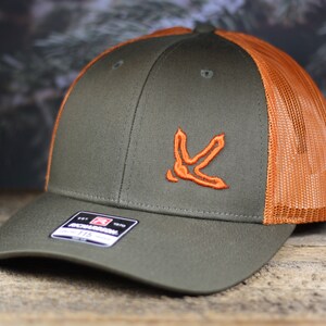 May include: A green and orange baseball cap with a brown bird track logo embroidered on the front. The cap has a mesh back.
