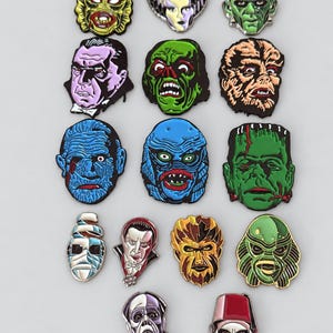 May include: A collection of fourteen enamel pins featuring stylized portraits of classic horror movie monsters and characters, including various green, blue, and purple hued creatures, vampires, and mummies, arranged in a grid on a plain background.