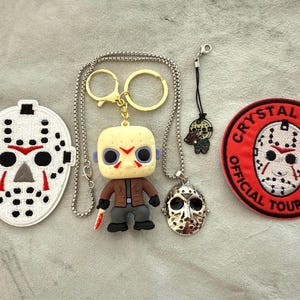 May include: Collection of horror-themed items, including a white hockey mask patch, a Funko Pop figure, a necklace with a mask pendant, a keychain, and a Crystal Lake patch. The items are arranged on a light gray surface.