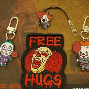 May include: A collection of horror-themed accessories, including two keychains with cartoon clown designs, a patch with the words "FREE HUGS", and a charm with a clown holding a red balloon. The items are in shades of red, black, white, and purple.