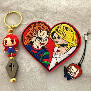 May include: A keychain featuring a small Chucky figure and a grey coffin charm, a heart-shaped embroidered patch depicting two characters with red and blonde hair, and a small Chucky character charm attached to a black cord with a metal clasp.