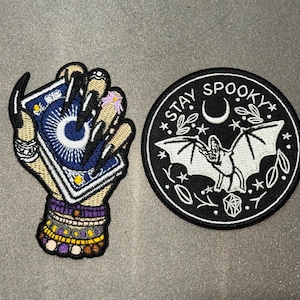 May include: Two embroidered patches. One patch depicts a hand with long black nails holding a blue tarot card with a white sunburst design, adorned with beaded bracelets. The other is a circular black patch featuring a white bat, a crescent moon, and the text STAY SPOOKY.