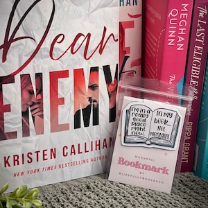 May include: A pink and blue magnetic bookmark with the text "I'm in a really good place right now. In my book... Not mentally."  The bookmark is shaped like an open book and has a white background. The text "Magnetic Bookmark by Blissfully Booked Co." is printed on the bottom of the bookmark.