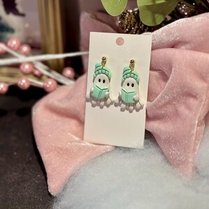 Pode incluir: Um par de brincos de novidade com pequenas figuras de bonecos de neve brancos com chapéus e livros verde-menta. Os brincos têm pinos dourados e são exibidos em um cartão branco. O fundo inclui tecido rosa e vegetação.