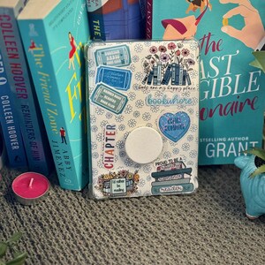 K&ouml;nnte beinhalten: Eine wei&szlig;e Handyh&uuml;lle mit Buchthema und bunten Aufklebern. Die Aufkleber zeigen B&uuml;cherstapel, Blumen und Text, der "B&uuml;cher sind mein gl&uuml;cklicher Ort" und "Bookwhore" sagt.