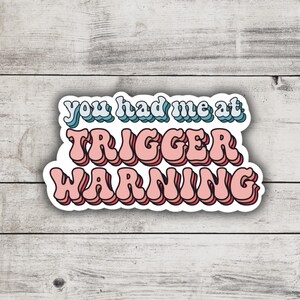 May include: A white sticker with the text "You had me at trigger warning" in pink and blue letters with a retro font.