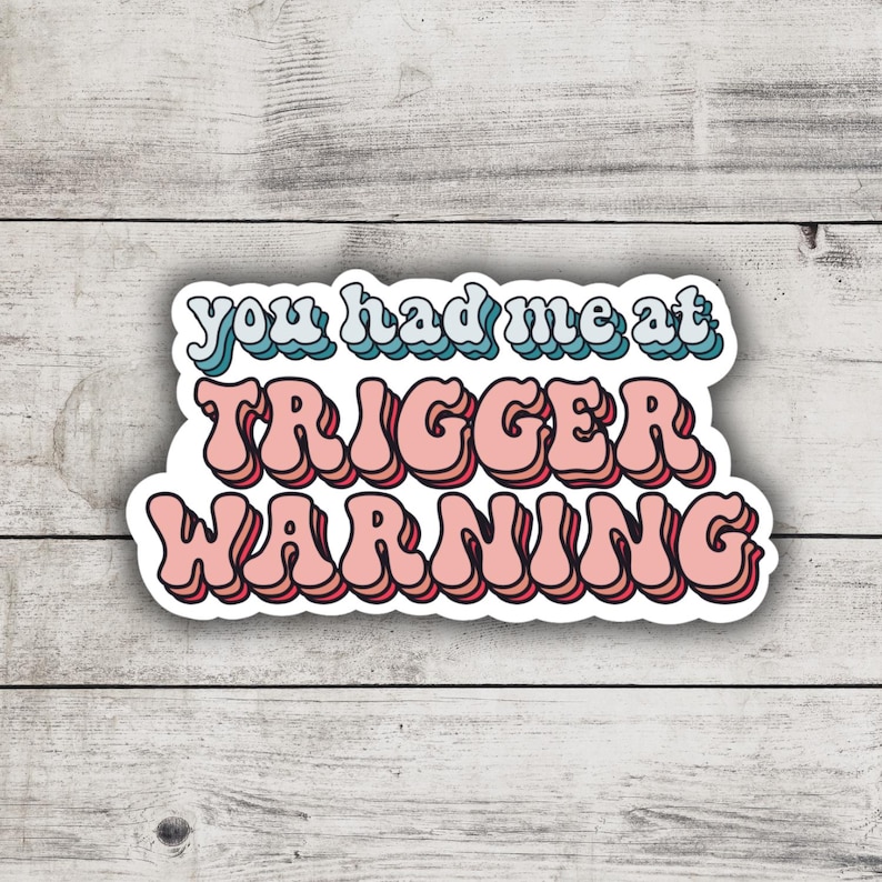 May include: A white sticker with the text "You had me at trigger warning" in pink and blue letters with a retro font.