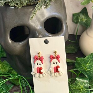 Peut inclure: Une paire de boucles d'oreilles fantômes faites à la main. Chaque boucle d'oreille représente un fantôme blanc avec un nœud rose, une écharpe rouge et rose et un clou doré. Les boucles d'oreilles sont présentées sur une carte blanche, avec un vase en forme de crâne en arrière-plan.