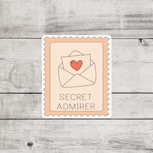 May include: A pink and white sticker with a cartoon illustration of a sealed envelope with a red heart inside. The text "SECRET ADMIRER" is printed below the envelope.