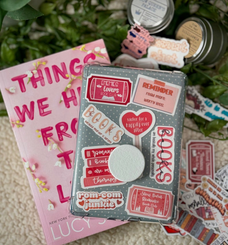 May include: A gray phone case with a variety of colorful stickers. The stickers feature book-themed quotes and illustrations, including "Enemies to Lovers Book Club", "Books because murder is wrong", "Rom-Com Junkie", and "Slow Burn Romance Book Club".