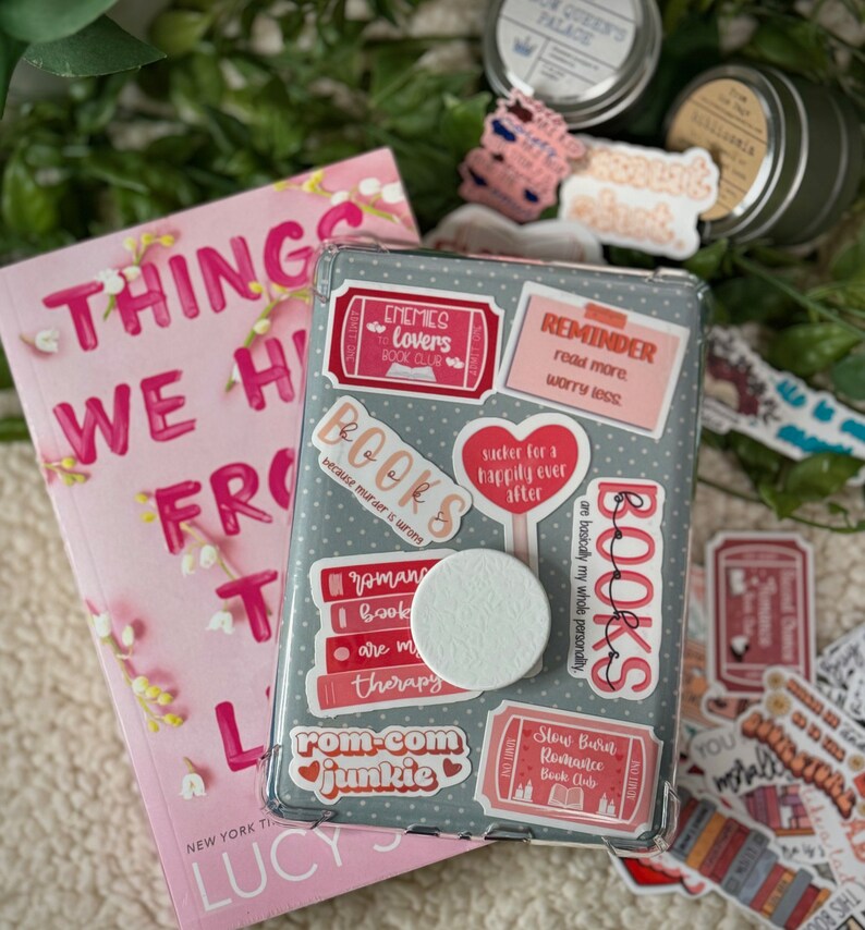 May include: A gray phone case with a variety of colorful stickers. The stickers feature book-themed quotes and illustrations, including "Enemies to Lovers Book Club", "Books because murder is wrong", "Rom-Com Junkie", and "Reminder: Read more, worry less".