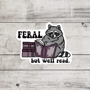 May include: A sticker with a cartoon raccoon reading a book. The raccoon is sitting on a stack of books. The text on the sticker reads "Feral but well read."