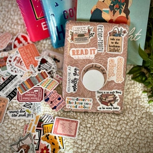 May include: A brown box with a variety of colorful stickers on it. The stickers feature book-related quotes and illustrations, including "Read it like a lady", "You had me at morally gray", "Take me to the bookstore", and "There is no such thing as too many books".