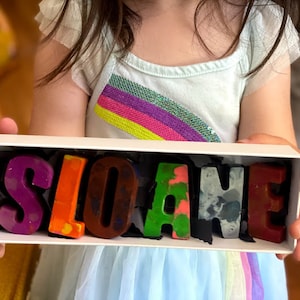 May include: A white box with the name "SLOANE" spelled out in colorful crayon letters. The letters are arranged in a row and are surrounded by black shredded paper.
