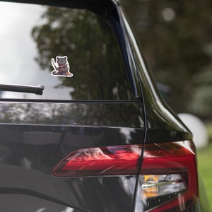May include: A black car with a sticker of a black cat wearing a red and gold outfit and holding a sword. The cat is looking to the left.